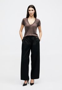 Femme debout, les mains dans les poches, portant un t-shirt marron à col en V, un pantalon noir à jambes larges et des chaussures à talons noires sur un fond uni.