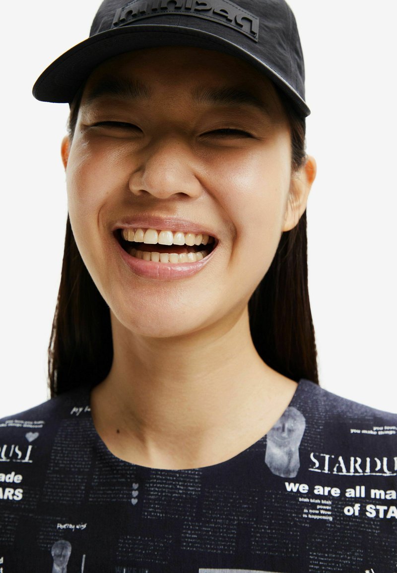 Jeune personne souriante avec des cheveux lisses et foncés, portant une casquette noire et un t-shirt sombre imprimé de texte et d'images blanches.