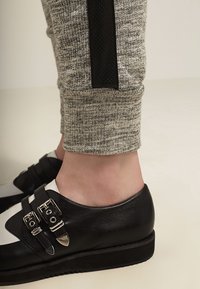 Grå teksturerede joggere med sort side-stribe, parret med sorte lædersko med to sølvspænder og en hvid detalje.