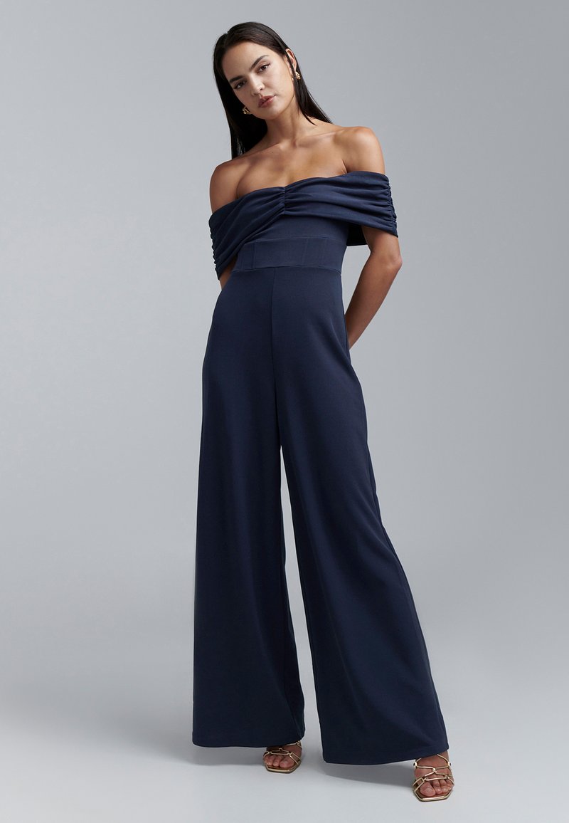 Tamsiai mėlynas kombinezonas be petnešėlių, su raukšlėtu liemeniu, platėjantys platūs kelnės ir minkšta medžiaga. Papildytas auksinėmis aukštakulnėmis su dirželiais.