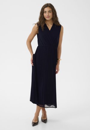 Kobieta o kręconych włosach, ubrana w granatową plisowaną sukienkę midi bez rękawów oraz czarne szpilki z ostrym noskiem, stoi z jedną ręką w kieszeni.