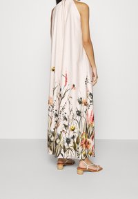 Mujer vistiendo un vestido maxi sin mangas de color rosa claro con un estampado floral colorido en el dobladillo, combinado con sandalias de tacón beige.