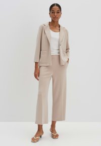 Blazer beige aderente con due tasche frontali, abbinato a pantaloni larghi coordinati e una maglietta bianca. Sandali slip-on con cinturini sottili.