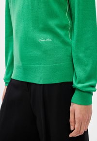 Πράσινο πλεκτό πουλόβερ με ριμπ μανσέτες και στρίφωμα, featuring λευκό κεντημένο λογότυπο στο μπροστινό μέρος. Συνδυασ�μένο με μαύρες παντελόνες.