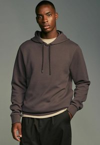 Brun struktureret hoodie med snoretræk, prydet med et subtilt diamantmønster, parret med en lysfarvet skjorte underneath.