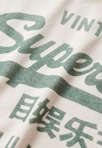 Witte katoenen t-shirt met grote, vervaagde groene typografie. De tekst bevat "Super" en "Vintage" met een zachte, gestructureerde uitstraling.