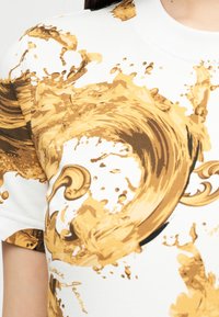 Κοντινό πλάνο άσπρου πουκαμίσου με στροβιλιζόμενο μοτίβο από βαφή σε χρυσό-καφέ αποτύπωμα στον ώμο και στο στήθος, που φοριέται από ένα άτομο.