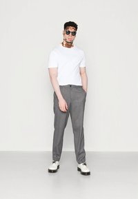 Valge lühikeste varrukatega t-särk, hallid kohandatud püksid ja valged kingad. Modell kannab päikeseprille, seisab ühevärvilise tausta ees.