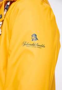 Mâneca unei jachete de ploaie galben strălucitor cu un logo negru care prezintă un bărbat cu barbă purtând o pălărie de ploaie și textul "Schmuddelwedda Wenn dat pladdert".
