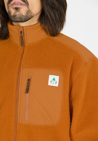 Veste en polaire orange avec un col haut, une poche poitrine zippée et un patch logo. La texture est douce et pelucheuse.