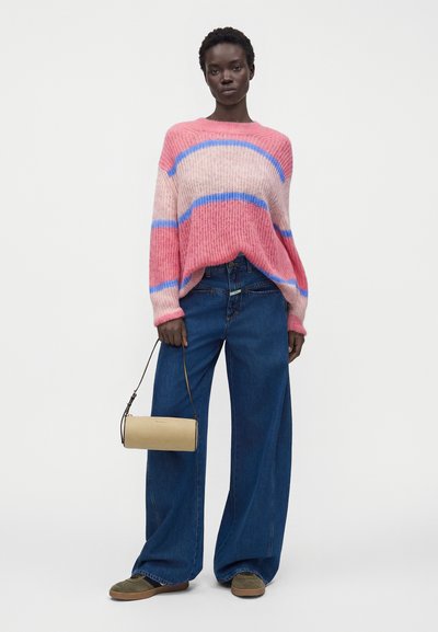 Femme portant un pull rayé rose et bleu, un jean bleu à jambes larges, des baskets vertes, tenant un petit sac à main beige par sa bandoulière.