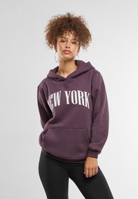 Ljubičasti dukserica s natpisom "NEW YORK" ispisanim bijelom bojom. Sadrži kengurua džep, podesivu kapu s vezicom i slobodan kroj. Uparen s crnim tajicama.