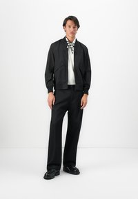 Man wearing black bomber jacket, black pants, black shoes, white shirt, and black-and-white patterned neck scarf, standing against white background.
