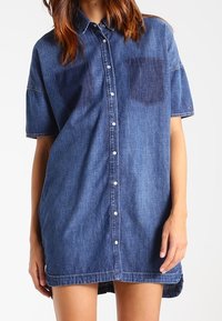 Denim-paitamekko lyhyillä hihoilla, vaaleansinistä kangasta, kaksi rintataskua, napitus edessä ja lievästi kaareva helma.