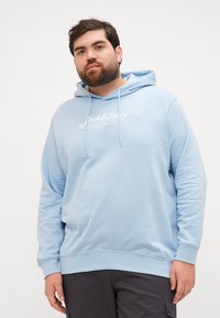 Světle modrá mikina s kapucí, která má přední klokaní kapsu, stahovací šňůrku a bílý logo značky. Vyrobena z měkké tkaniny s žebrovanými manžetami a spodním lemem.