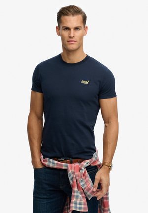 Uomo con t-shirt blu navy con piccolo logo, camicia a quadri annodata in vita, jeans scuri e orologio da polso su sfondo semplice.