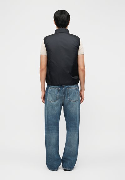 Άνδρας που στέκεται με την πλάτη, φοράει μαύρο αμάνικο γιλέκο με επένδυση πάνω από μπεζ μπλουζάκι και άνετο μπλε τζιν, σε απλό λευκό φόντο.