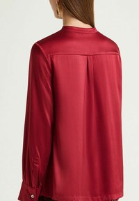 Rote Satinbluse mit langen Ärmeln, einem Mandarinkragen und Knopfdetails an den Manschetten, mit einer Falte auf der Rückseite für zusätzliche Details.