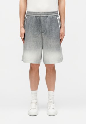 Homme portant un short dégradé du gris au blanc longueur genou, des chaussettes blanches, des baskets blanches et une chemise blanche, debout les mains relâchées.