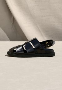 Sandale en cuir noir pour homme avec larges brides, orteil ouvert, bride arrière avec boucle dorée, vue de côté sur une surface claire.
