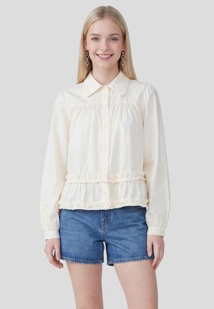 Młoda kobieta z długimi blond włosami, nosząca zwisające kolczyki w kształcie serca, kremową bluzkę z długim rękawem i falbankami oraz niebieskie jeansowe szorty, uśmiechająca się.