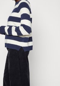 Pull en tricot bleu marine et crème à rayures, à manches longues, avec des bords côtelés et un ourlet asymétrique. Assorti avec un pantalon en velours côtelé foncé.