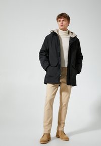 Abrigo negro con capucha y forro suave color beige, combinado con un cuello de tortuga beige claro y pantalones beige. Botas marrones de tobillo completan el look.