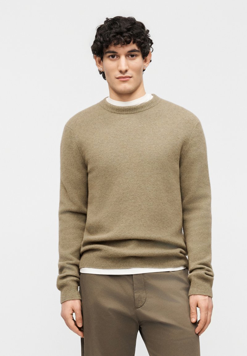 Νέος άνδρας με σκούρα σγουρά μαλλιά, φορώντας μπ εζ πουλόβερ με κυκλικό λαιμόκοψη πάνω από άσπρο πουκάμισο και καφέ παντελόνι, στέκεται μπροστά σε απλό φωτεινό φόντο.