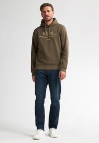 Olivově zelená mikina s kapucí a tištěnými grafikami, kombinovaná s tmavě modrými džíny a bílými teniskami. Hladká textura, volný střih.