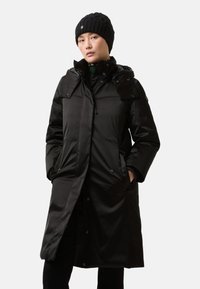 Zwarte lange pufferjas met een capuchon, voorzien van een ritssluiting, drukknoopsluiting, klepzakken en geribbelde manchetten. Draagt een zwarte gebreide muts.