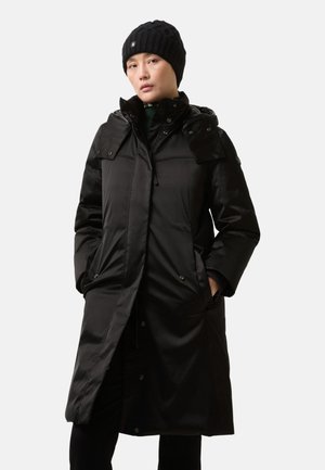 Manteau puffer long noir avec capuche, dotée d'une fermeture éclair, de boutons-pression, de poches à rabat et de poignets côtelés. Porté avec un bonnet en tricot noir.