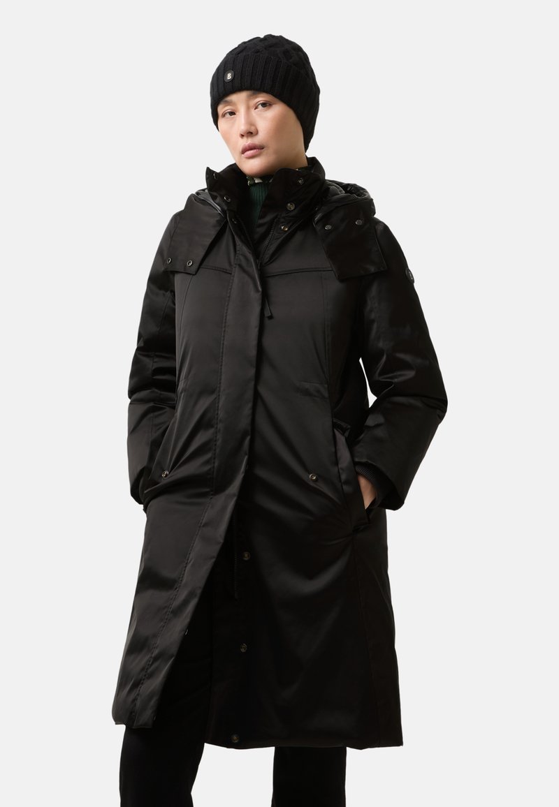 Zwarte lange pufferjas met een capuchon, voorzien van een ritssluiting, drukknoopsluiting, klepzakken en geribbelde manchetten. Draagt een zwarte gebreide muts.