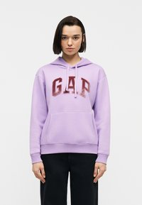 Νεαρό άτομο με κοντά σκούρα μαλλιά, που φοράει λιλά φούτερ με κόκκινα γράμματα "GAP" και μαύρο παντελόνι, στ έκεται μπροστά σε απλό λευκό φόντο.