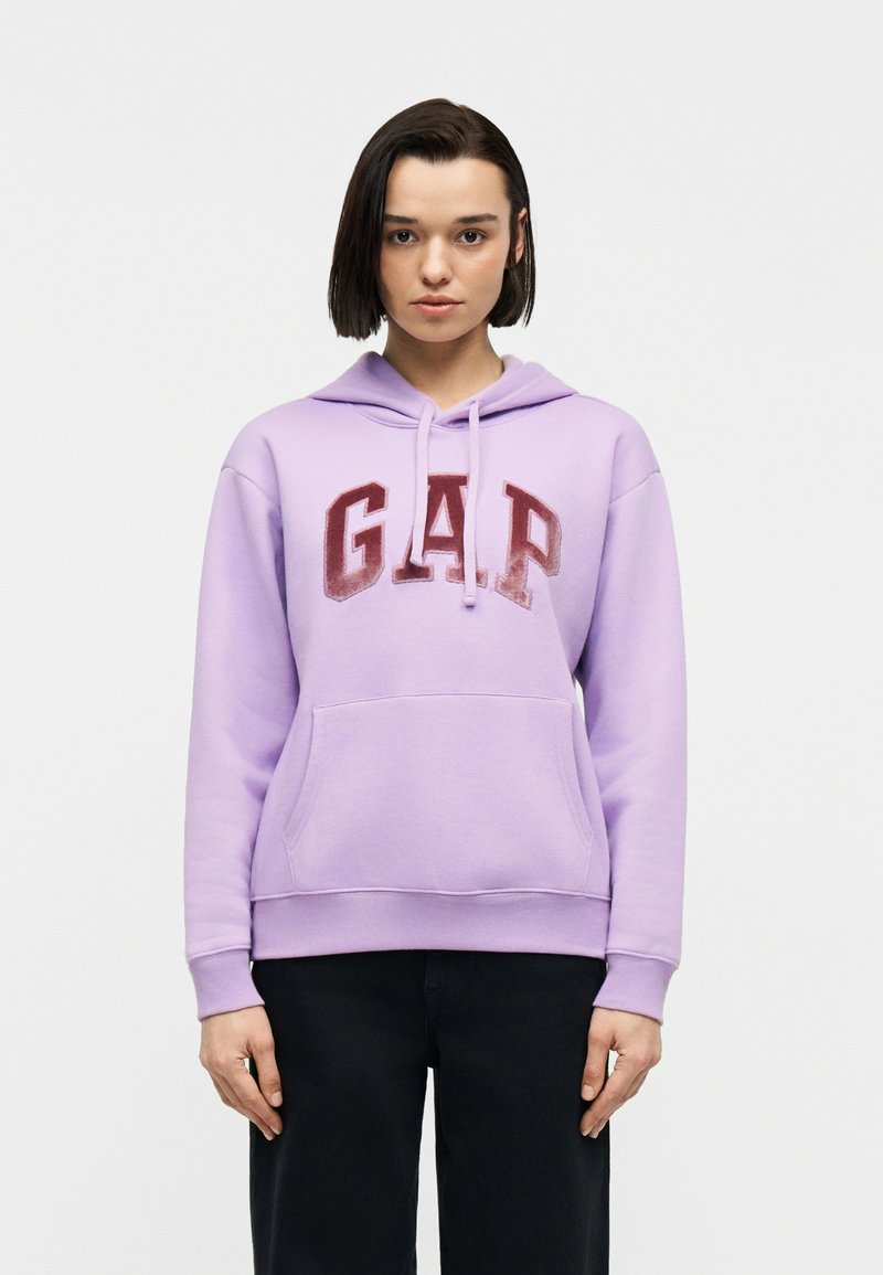 Νεαρό άτομο με κοντά σκούρα μαλλιά, που φοράει λιλά φούτερ με κόκκινα γράμματα "GAP" και μαύρο παντελόνι, στέκεται μπροστά σε απλό λευκό φόντο.