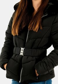 Chaqueta negra acolchada con acabado texturizado, que cuenta con capucha de piel sintética y un cinturón negro con tachuelas como detalle. Cierre de cremallera y bolsillos laterales.