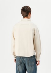 Beige paitapusero, jossa on kaulus ja pitkät hihat, rento istuvuus ja helma, joka laskeutuu hieman taakse.