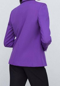 Blazer violet sur mesure avec un col cranté, une texture lisse et une forme structurée. Les manches présentent un léger froncé aux épaules.