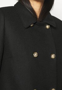 Abrigo negro de doble botonadura con una tela texturizada, un amplio cuello y botones decorativos en tono dorado con superficie texturizada.