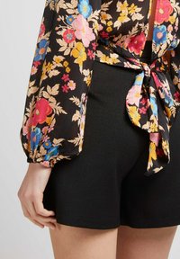 Blouse florale avec des manches longues bouffantes en tissu noir, ornée de fleurs colorées en rose, bleu et jaune. Associée à un short noir ajusté.