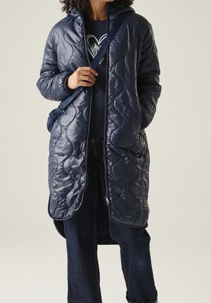 Osoba ubrana w długą granatową pikowaną kurtkę, ciemne dżinsy i granatową koszulę z wzorem przedstawiającym górskie serce, niosąca granatową torbę przewieszoną przez ramię.