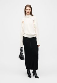 Γυναίκα που φοράει λευκό πουλόβερ με ανοίγματα, μακριά μαύρη φούστα, μαύρες μπότες με ψηλό τακούνι, κρατώντας μαύρη τσάντα χειρός μπροστά σε απλό φόντο.