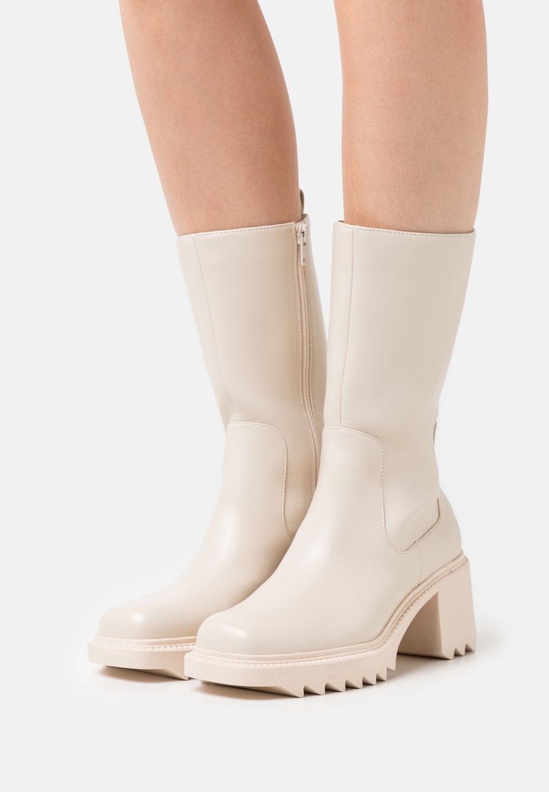 Bottes mi-mollet couleur crème avec talons blocs épais, fermetures éclair latérales et semelles striées, portées par une personne aux jambes nues.