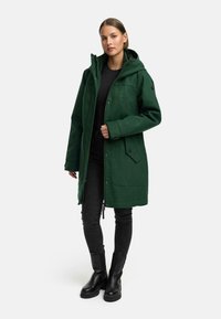 Groene wolmix parka met een capuchon, drukknoopsluiting en twee zijzakken. Gecombineerd met een zwarte top, jeans en zwarte enkellaarzen.