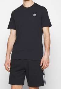 Muž oblečený v černém tričku Adidas a černých šortkách s bílými logy a pruhy Adidas na jednobarevném pozadí.