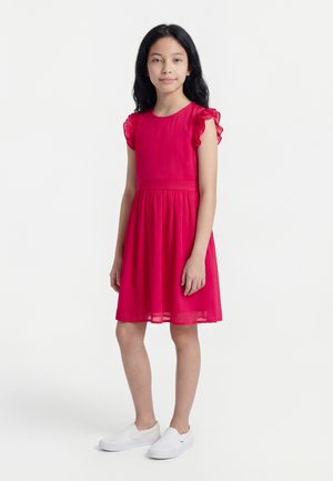 Dziewczyna z długimi czarnymi włosami, ubraną w czerwoną sukienkę bez rękawów z falbanami na ramionach oraz białe buty wsuwane, stojąca na białym tle.