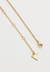 Goldfarbene Kette mit einem dreieckigen Anhänger, der mit der Gravur "GUESS" versehen ist, gesichert mit einem Karabinerverschluss und runden Gliedern.
