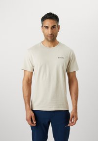 T-shirt en coton beige avec col rond et manches courtes. Présente un petit logo noir sur la poitrine gauche. Texture lisse, coupe décontractée.