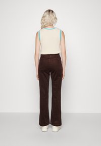 Femme avec des cheveux blonds ondulés portant un haut sans manches crème avec des bords bleus, un pantalon en velours côtelé marron et des chaussures à plateforme blanches, de dos.