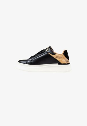 Zapatilla negra de cuero con un acabado brillante, suela blanca texturizada y un acento en el talón de color marrón con patrón. Presenta cordones negros y ojetes dorados.