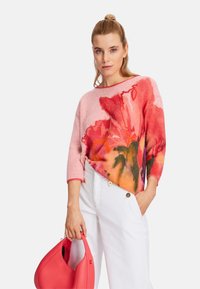 Knit pullover in pink with vibrant floral print, rounded neckline, and three-quarter sleeves. Paired with white wide-leg pants and a red bag.